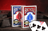 Two Bicycle double-faced playing card decks standing upright on dark stones, with and example of a double faced  against a warm red and dark background.