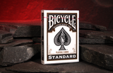 A boxed deck of Black Bicycle Standard playing cards standing upright on dark slate stones against a glowing red  cave background.