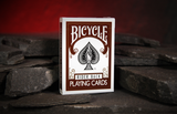 A box of Bicycle Rider Back Playing Cards standing upright on dark slate pieces against a warm red cave background.