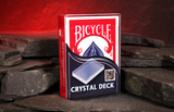 A boxed deck of Crystal Deck Magic Trick Bicycle Playing Cards standing upright on dark stacked stones, with a glowing red cave background.