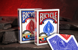 Two Bicycle Double Red & Blue Backed Playing Card Boxes standing upright on a dark stone surface. A single card lying in front showing the blue and red back design against a warm red cave background.