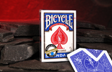 A box of Bicycle Double Backed Magic Cards stands upright on dark slate stones, with one double blue-backed playing card placed in front on the same surface against a warm red glowing background.