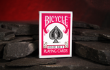 A boxed deck of  Fuchsia Bicycle Rider Back Playing Cards stands upright on dark slate stones against a warm red cave background.