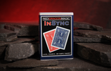 A boxed deck of cards standing upright on dark stone blocks against a  warm red glowing backdrop. The box reads REDDRAGONMAGIC INSYNC and shows red and blue card backs on the front.