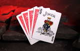 A fan of playing cards, with the top card showing a Joker illustration, resting on dark stone blocks against a glowing red cave background.