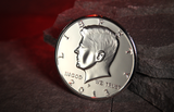 A silver jumbo half dollar featuring a profile portrait of John F. Kennedy, with the text 'LIBERTY used for coin magic. The coin is resting in a cave with a glowing red background.