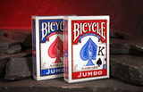 Two Bicycle jumbo playing card decks standing upright on dark stones against a warm red cave background. One deck has a red box and blue lettering, and the other is a blue box and red lettering.