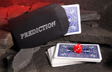 A black envelope labeled 'PREDICTION' with a blue-backed playing card partially pulled out, alongside a small stack of matching blue-backed cards and a red die, arranged on dark rocks with a glowing red background.