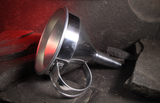 A polished stainless steel funnel with a loop handle, resting on dark textured stone against a glowing red cave background.