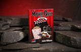 A boxed card game called "Movie Quiz" by Jamie Williams stands upright on dark slate stones against a warm red cave backdrop. The box is red with black, white, featuring movie-themed graphics including a clapperboard, popcorn bucket, film reel, and 3D glasses.
