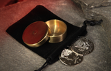 A brass coloured Okito Box for magic with its lid removed, resting on a black velvet drawstring pouch next to two replica silver half dollar palming coins on a gray stone surface with a glowing red cave background.