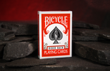A box of Bicycle Rider Back playing cards standing upright on dark slate stones against a warm red background.