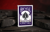A box of Bicycle Rider Back Playing Cards standing upright on dark slate stones against a warm red background.