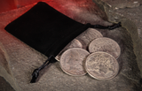 Five Replica Morgan Silver Dollars for Magic and Palming next to a black felt back resting on a cave floor with a glowing red background. 