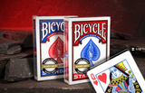 Two Bicycle short deck playing card boxes standing upright on a dark stone surface, with one red-suited box in front of a blue-suited box and a single 1/16" short Queen of Hearts card placed in front. The background is a dark warm red cave backdrop.
