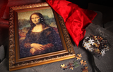 A framed jigsaw puzzle of the Mona Lisa, partially assembled and displayed on a cave floor with loose puzzle pieces and a bag of pieces beside it. A red satin silk cloth is draped behind the frame.