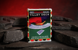 A boxed deck of cards standing upright on dark stone blocks against a warm red glow. The card box reads 'RED DRAGON MAGIC TILLEY DECK' and shows a green baise with a red-and-black jester's hat and an illustration of fanned cards.