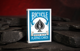 A deck of Turquoise Bicycle Playing Cards Rider Back on a stone floor with a warm red glow on the back cave wall. 
