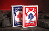 Two Bicycle Ultimate Left handed Playing Cards decks standing upright on dark slate stones against a warm red cave background.