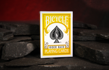 A deck of Yellow Rider Backed Bicycle Playing Cards on a dar slate floor with a warm red glowing cave background